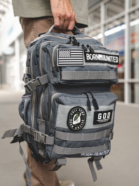 Someone holds the Pilot Gray Tac-Pac - 25L by Born United Private Label, adorned with patches like an American flag, BORN UNITED, and GOD. The scene is outdoors and the person wears brown pants.