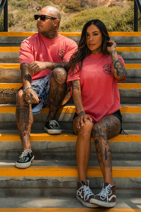A tattooed man and woman sit on outdoor stairs amid greenery, both in pink unisex Relentless Betrayal shirts—he sports the Into the Storm Premium Watermelon T-Shirt. Relaxed and stylish in shorts, they enjoy each others company.