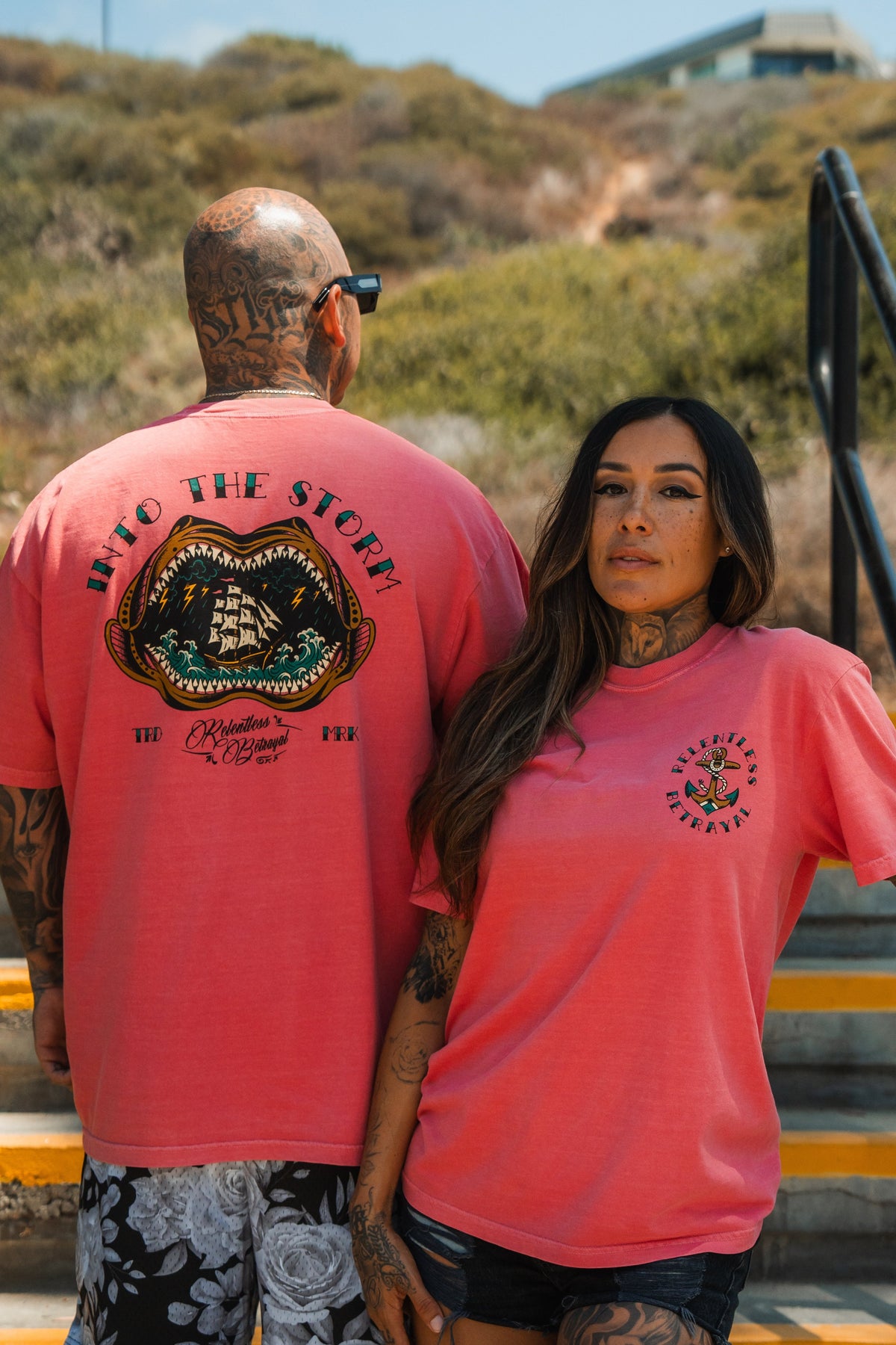 A tattooed man and woman stand on outdoor steps, both wearing Relentless Betrayals unisex Into the Storm Premium Watermelon T-Shirts; his shows a ship with “Into the Storm,” hers has a small chest with “Chivalrous Behavior.” Bushes and a building are behind them.