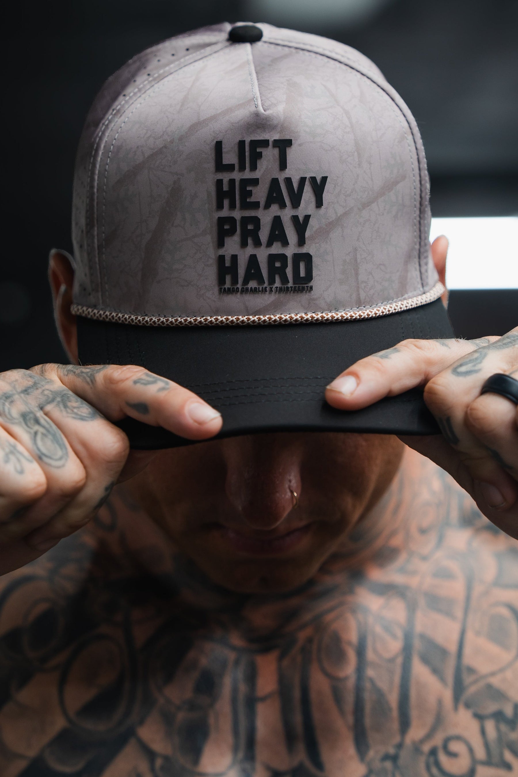 A tattooed individual holds the brim of Tango Charlie Apparel’s Lift Heavy. Pray Hard. Deadwood Camo Hydro Snapback Hat—an ultra-durable, sweat-resistant cap designed for those who push their limits.