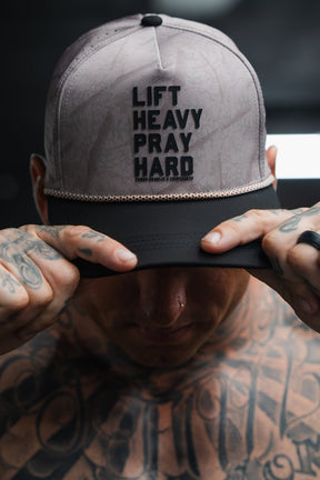 A tattooed individual holds the brim of Tango Charlie Apparel’s Lift Heavy. Pray Hard. Deadwood Camo Hydro Snapback Hat—an ultra-durable, sweat-resistant cap designed for those who push their limits.