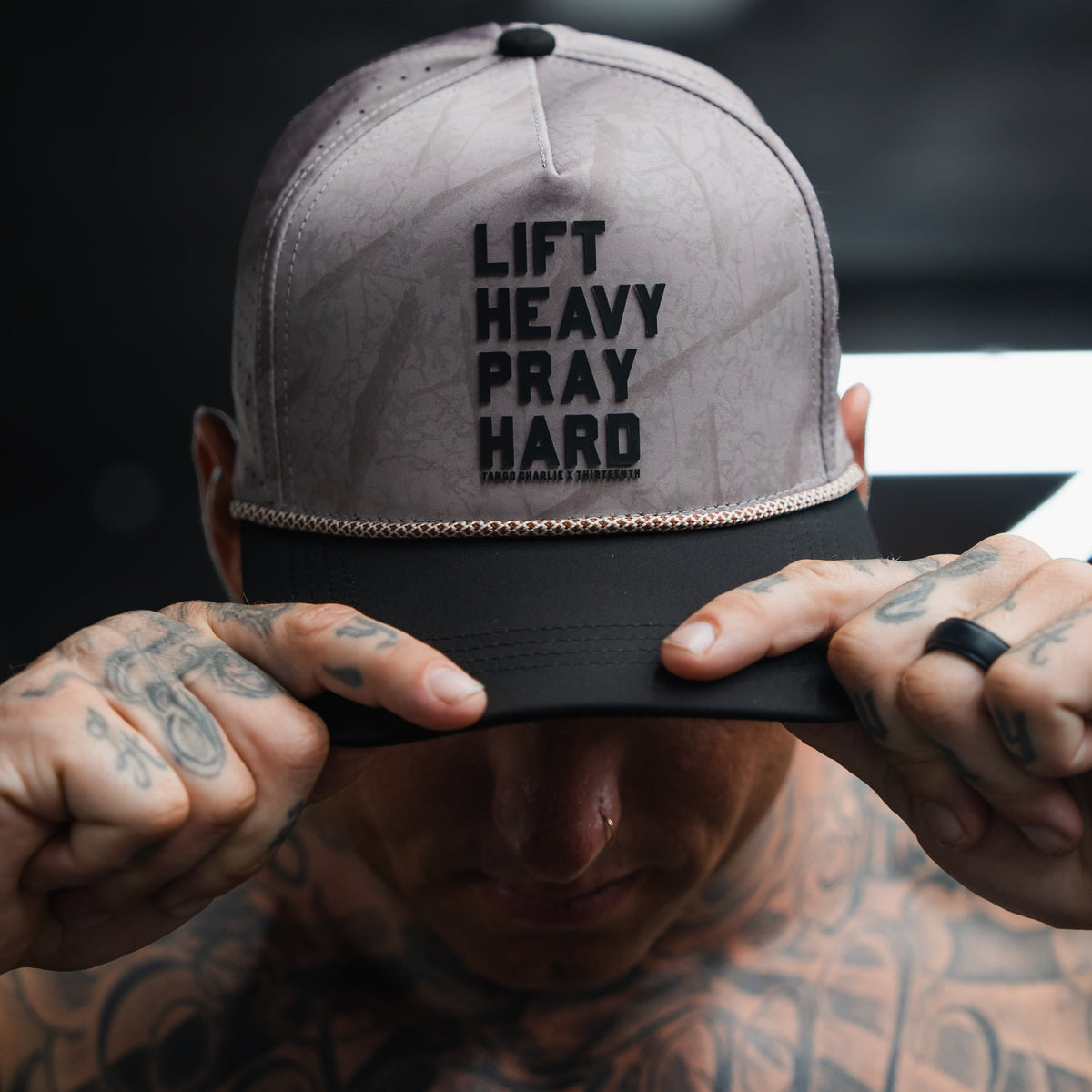 A tattooed individual adjusts a Tango Charlie Apparel Lift Heavy. Pray Hard. Deadwood Camo Hydro Snapback Hat. The background is blurred and the lighting is dim.