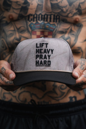 A shirtless, tattooed person holds a gray and black Tango Charlie Apparel “Lift Heavy. Pray Hard. Deadwood Camo Hydro” snapback hat with the words “LIFT HEAVY PRAY HARD” printed on the front.