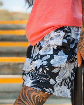 A person in Relentless Betrayals Premium Mesh Shorts | 5- Beauty in Darkness with a floral pattern and a pink shirt stands by yellow stairs, showing off detailed black tattoos on their arm and leg.