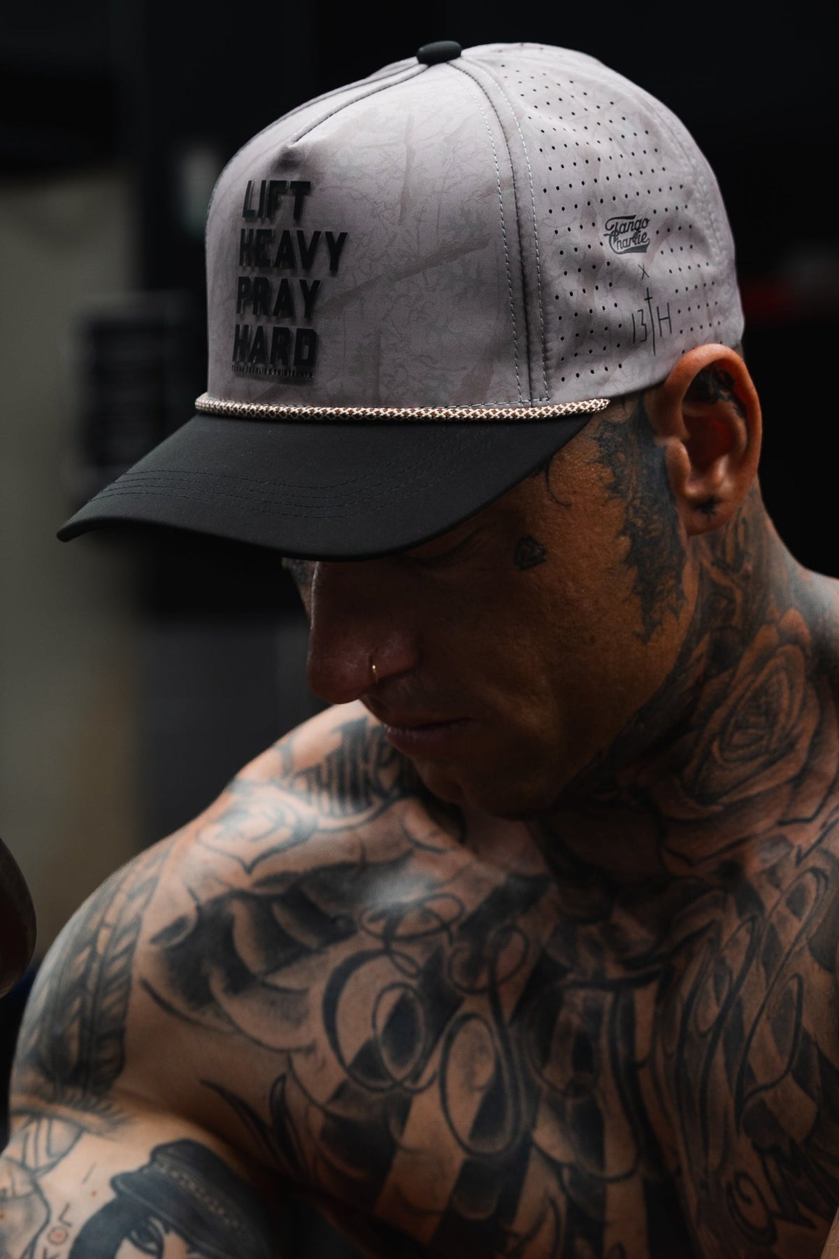 A tattooed individual wearing the Lift Heavy. Pray Hard. Deadwood Camo Hydro Snapback Hat by Tango Charlie Apparel in grey, gazing downward in a dimly lit environment.