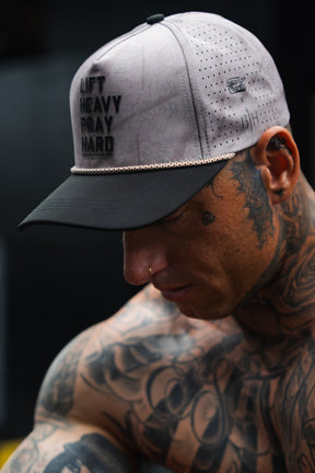 A tattooed man wears the Lift Heavy. Pray Hard. Deadwood Camo Hydro Snapback Hat by Tango Charlie Apparel, featuring sweat-resistant performance and bold style for dedicated lifters; his gray and black hat is visible as he looks down with his face partially in view.