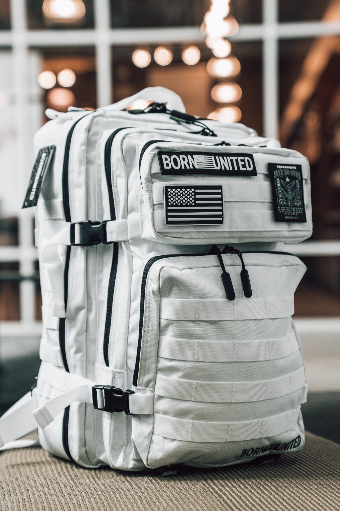 The Storm Trooper Tac-Pac - 45L by Born United Private Label, a white tactical backpack with black zippers and BORN UNITED and American flag patches, sits on a beige surface indoors with blurred lights and windows behind.