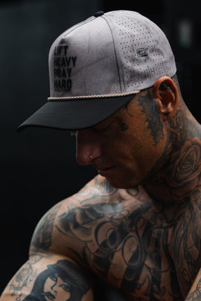 A tattooed individual wearing the Lift Heavy. Pray Hard. Deadwood Camo Hydro Snapback Hat by Tango Charlie Apparel gazes downward in a dimly lit setting.