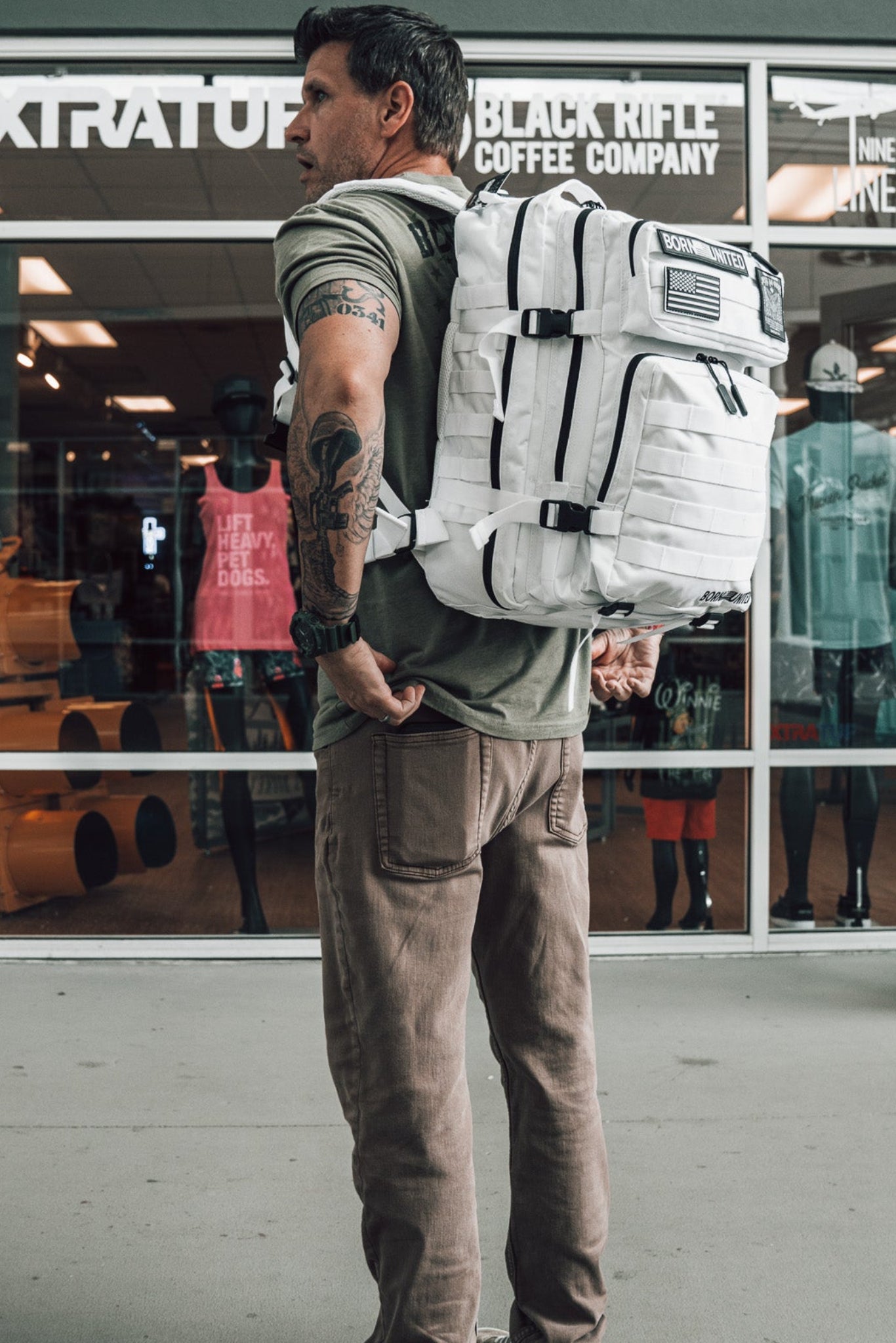 A tattooed man with a white backpack stands outside a store, wearing a green t-shirt and brown pants. Inside, Born United Private Label’s Storm Trooper Tac-Pac - 45L is on display among clothing and other items.