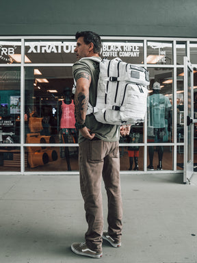 A tattooed man with a white backpack stands outside a store, wearing a green t-shirt and brown pants. Inside, Born United Private Label’s Storm Trooper Tac-Pac - 45L is on display among clothing and other items.