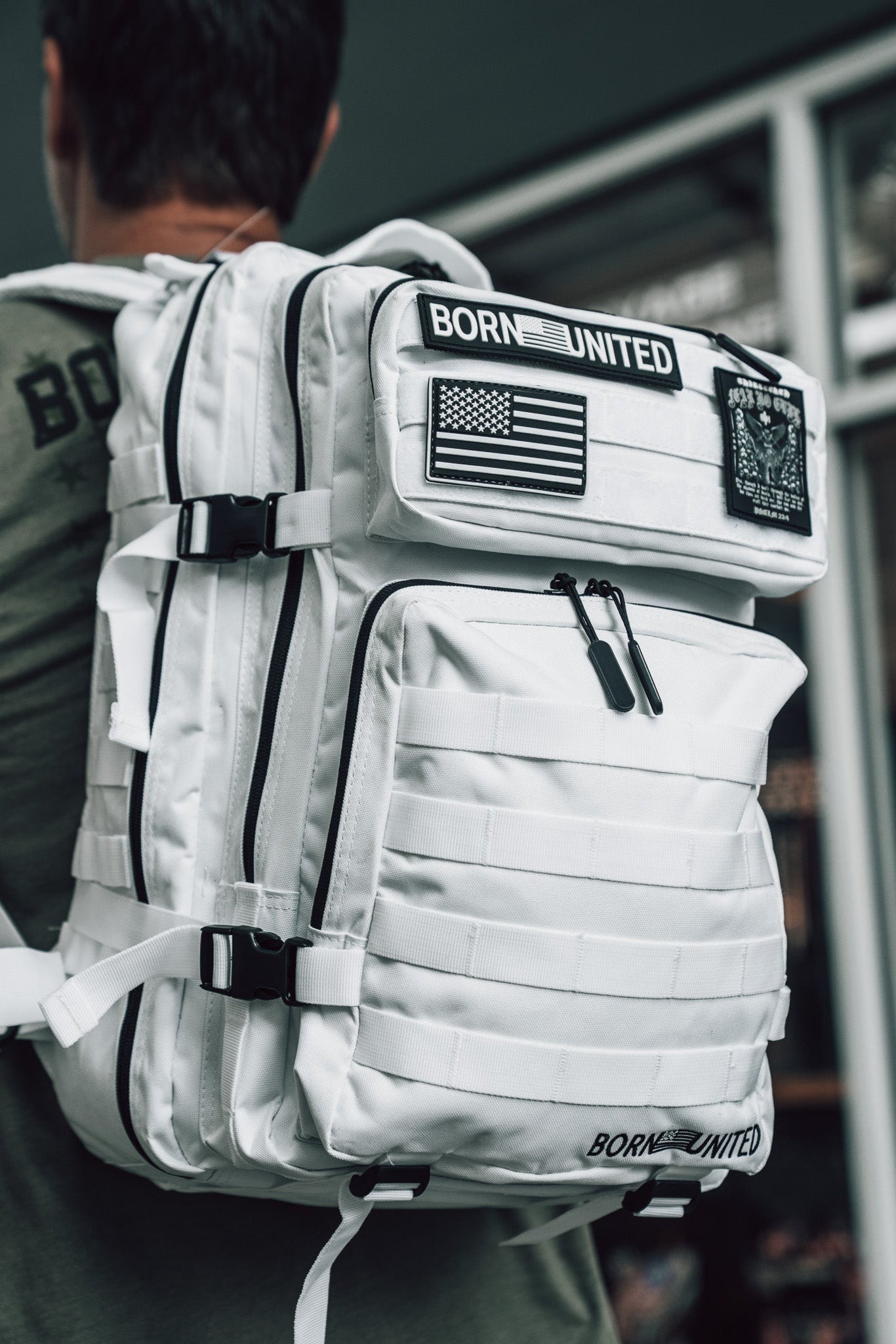 A person in an olive-green shirt carries the Born United Private Label Storm Trooper Tac-Pac - 45L, a large white tactical backpack with black zippers and patches including BORN UNITED, an American flag, and a subtle Storm Trooper helmet; background blurred.