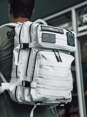 A person in an olive-green shirt carries the Born United Private Label Storm Trooper Tac-Pac - 45L, a large white tactical backpack with black zippers and patches including BORN UNITED, an American flag, and a subtle Storm Trooper helmet; background blurred.