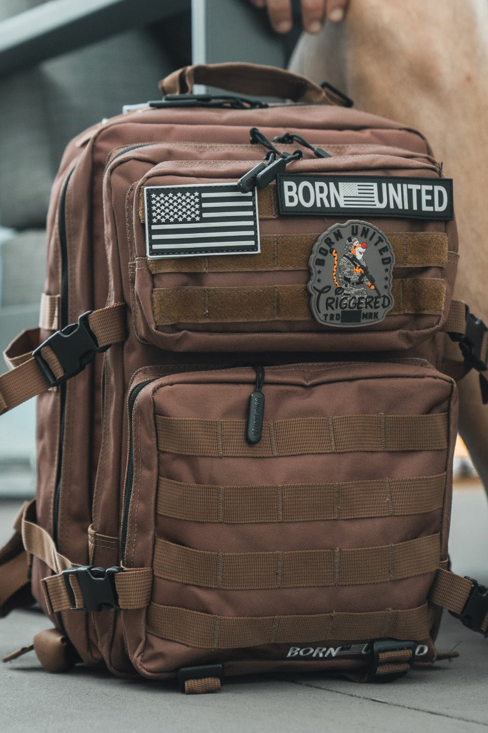 A Chocolate Tac-Pac - 25L by Born United Private Label, TSA-approved tactical backpack with multiple compartments and American flag, BORN UNITED, and TRIGGERED patches. Part of a person’s leg and shoe are visible beside the bag.