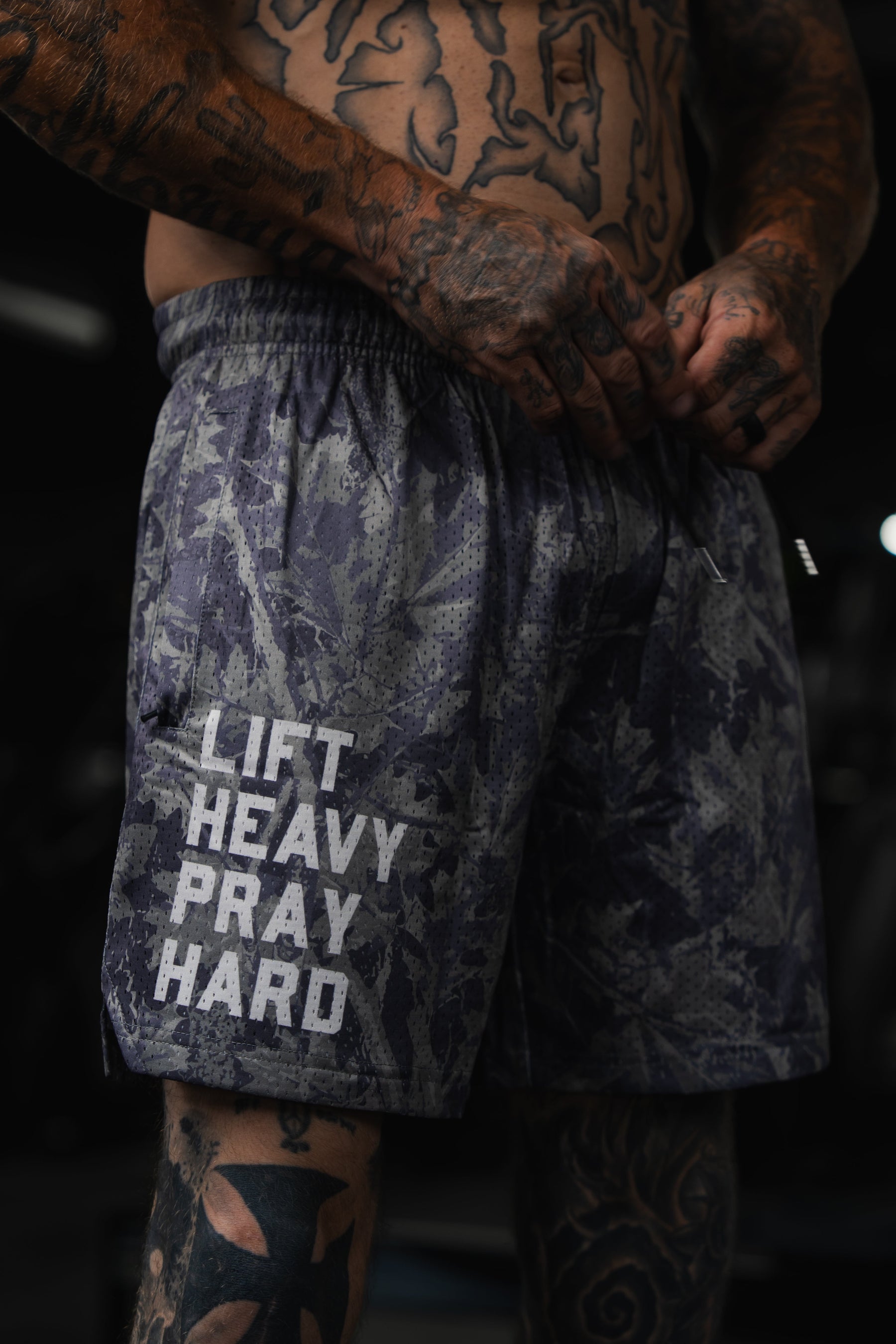 A tattooed person adjusts their Tango Charlie Apparel Men's Mesh Lifestyle Shorts | 5.5" - Lift Heavy Pray Hard Deadwood, featuring the text Lift Heavy Pray Hard printed on the left leg.