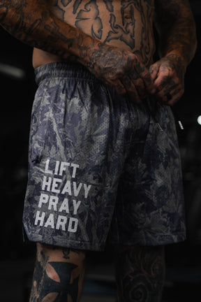 A tattooed person adjusts their Tango Charlie Apparel Men's Mesh Lifestyle Shorts | 5.5" - Lift Heavy Pray Hard Deadwood, featuring the text Lift Heavy Pray Hard printed on the left leg.