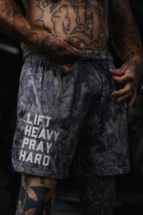 A tattooed individual stands in gym attire, sporting Tango Charlie Apparel's Men's Mesh Lifestyle Shorts (5.5") featuring "LIFT HEAVY PRAY HARD" in white text.