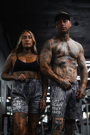 Two tattooed individuals stand side by side in a gym, both dressed in athletic wear. The man sports the Men's Mesh Lifestyle Shorts | 5.5" - Lift Heavy Pray Hard Deadwood from Tango Charlie Apparel, with the bold “LIFT HEAVY PRAY HARD” statement emblazoned on his shorts. Both face the camera with confidence, embodying a dedicated fitness lifestyle.