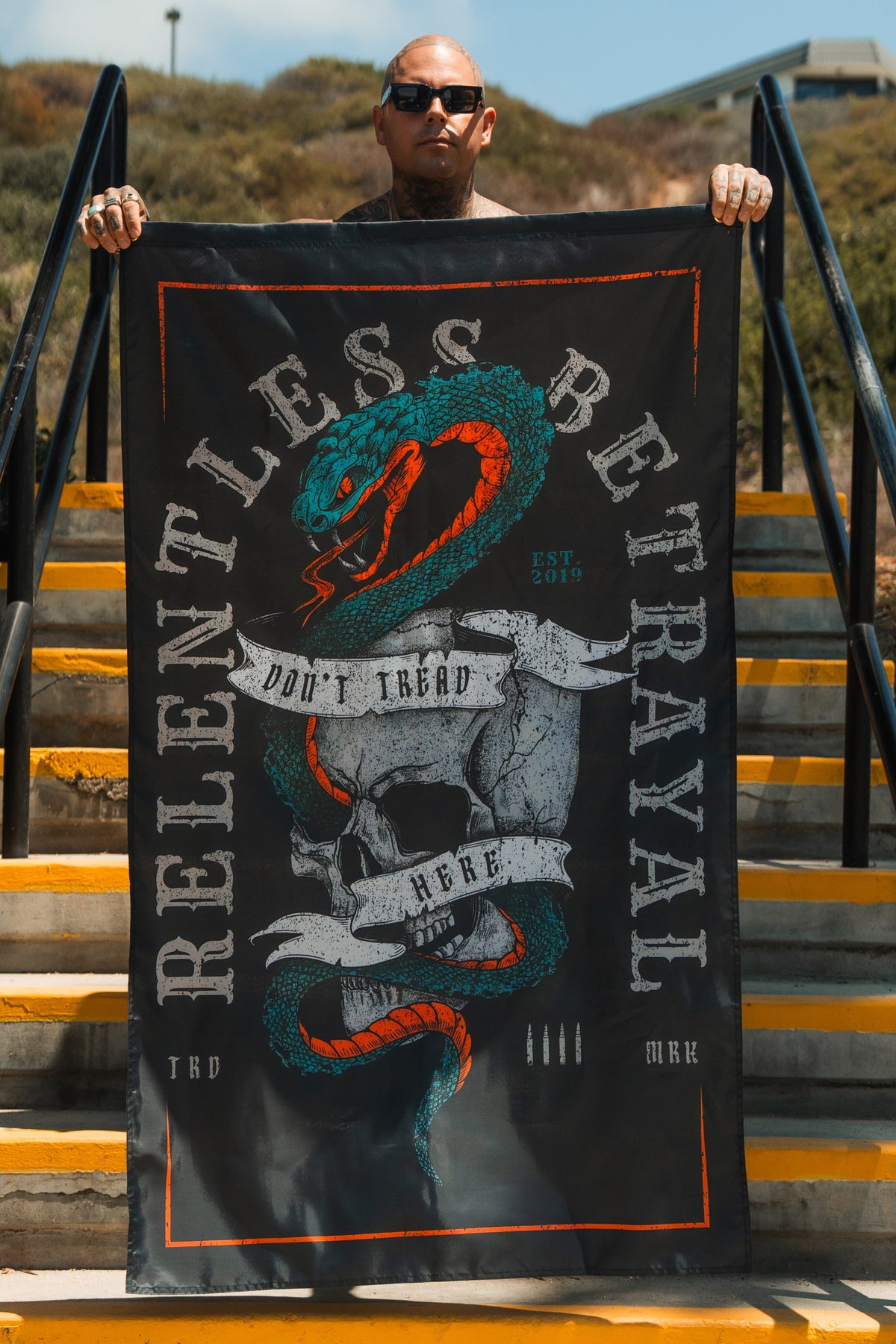Wearing sunglasses, a person stands on outdoor stairs holding the Dont Tread Here Flag by Relentless Betrayal, which displays a coiled snake, two skulls, and the bold phrases RELENTLESS BETRAYAL and DONT TREAD HERE.