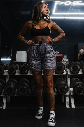 A woman with tattoos stands in front of a rack of dumbbells at a gym, wearing a black sports bra, Tango Charlie Apparel's Men's Mesh Lifestyle Shorts (5.5" - Lift Heavy Pray Hard Deadwood) with an eye-catching pattern, and sneakers.