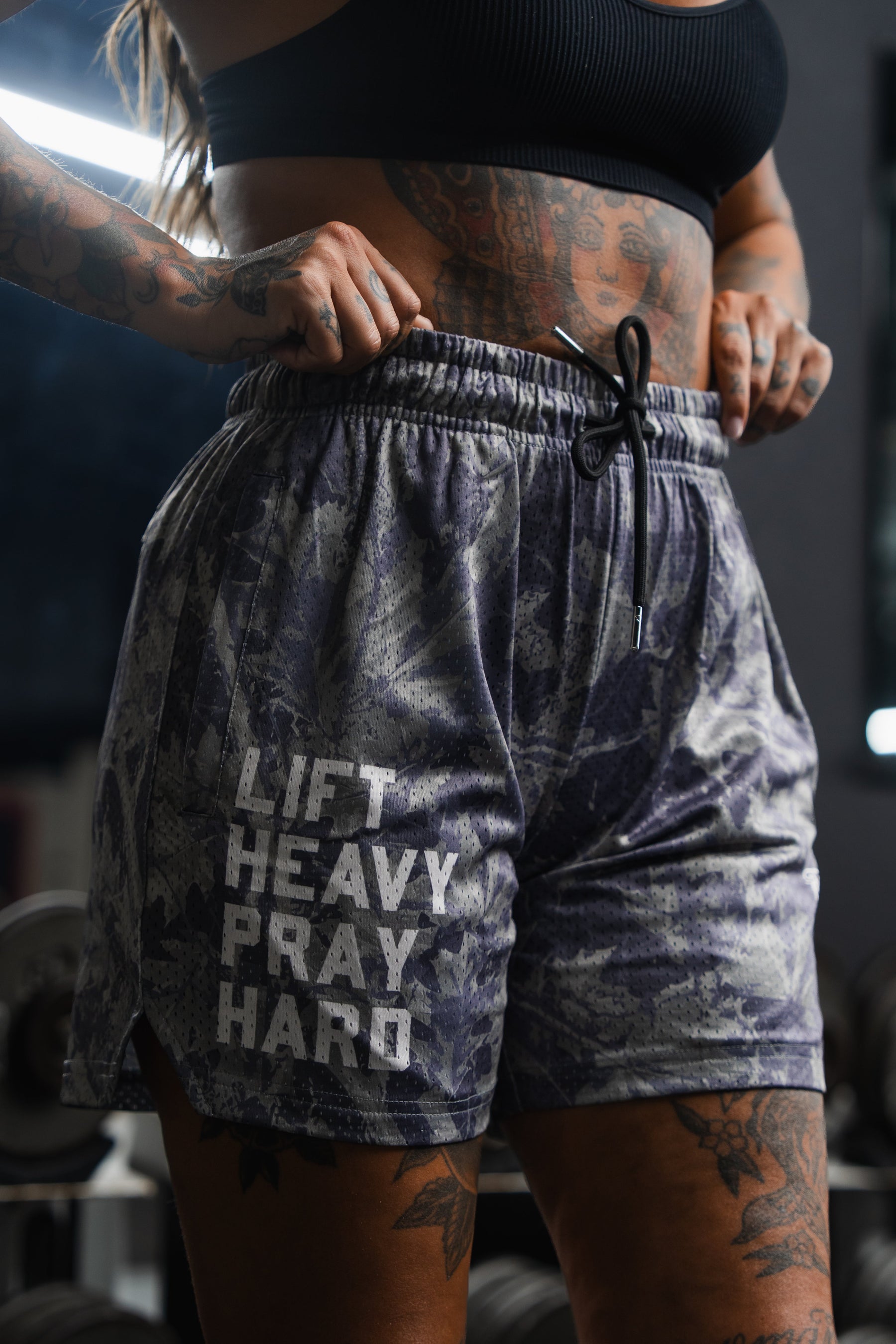 A tattooed individual is wearing Tango Charlie Apparel's Men's Mesh Lifestyle Shorts (5.5" - Lift Heavy Pray Hard Deadwood), standing in a gym with dumbbells in the background.