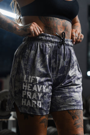 A tattooed individual is wearing Tango Charlie Apparel's Men's Mesh Lifestyle Shorts (5.5" - Lift Heavy Pray Hard Deadwood), standing in a gym with dumbbells in the background.