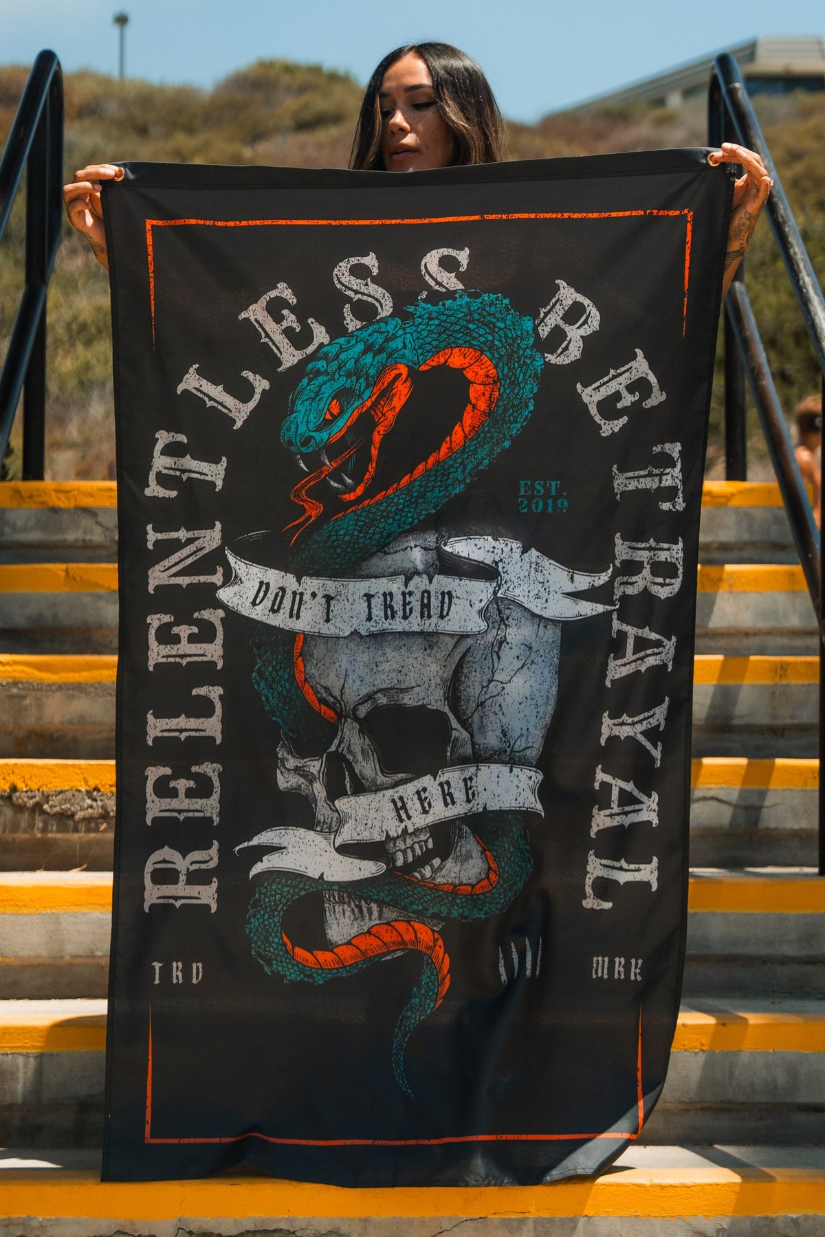 A woman stands on yellow steps holding the Dont Tread Here Flag by Relentless Betrayal, featuring a black banner with a coiled snake and orange, teal, and white accents for a gritty, bold look inspired by youth culture.
