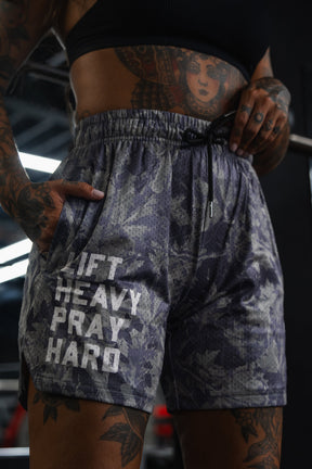 A person wearing Tango Charlie Apparel's Men's Mesh Lifestyle Shorts (5.5" - Lift Heavy Pray Hard Deadwood) stands with hands in pockets, showcasing visible tattoos on their arms and torso.