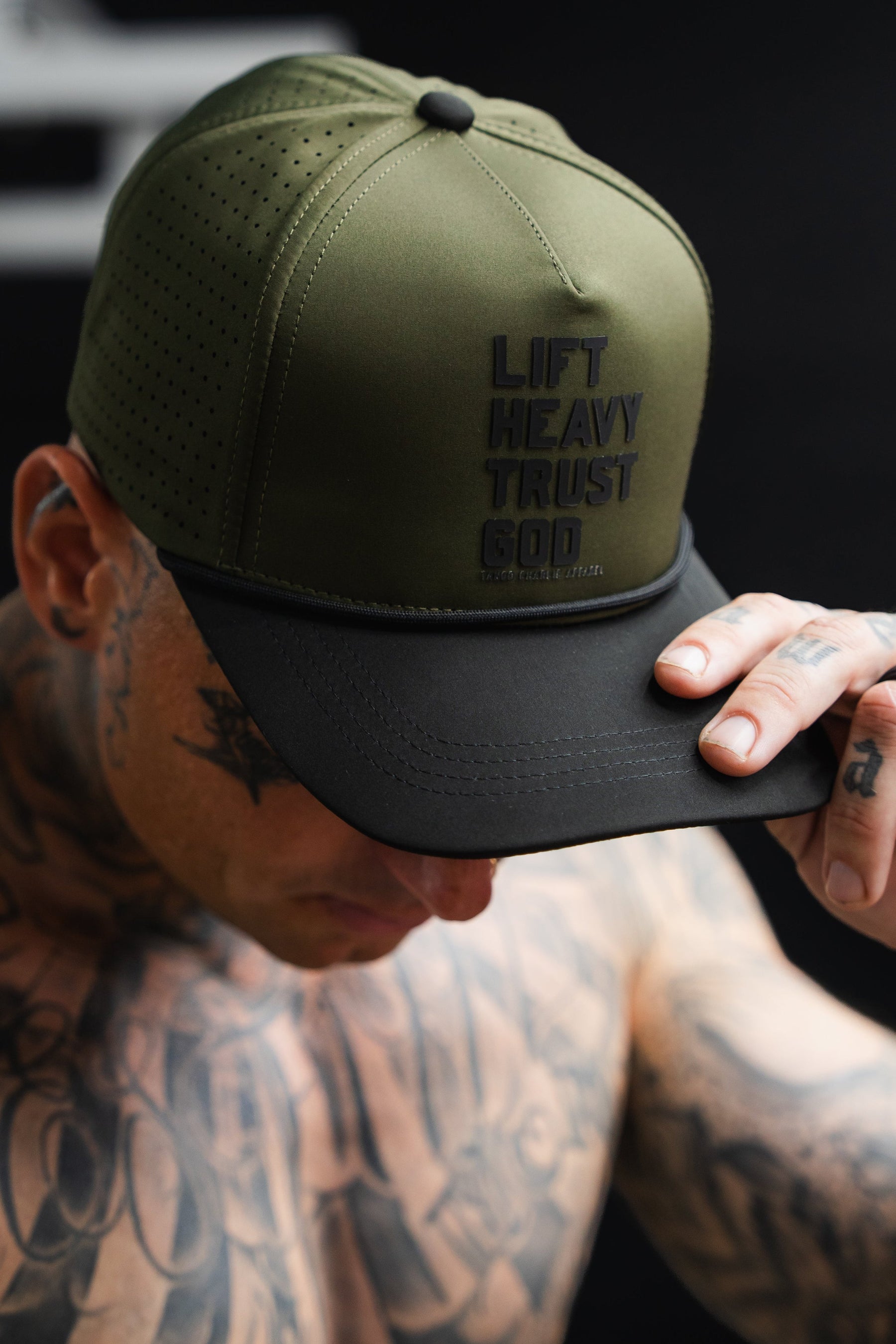 A tattooed individual wears the Tango Charlie Apparel Lift Heavy. Trust God. Hydro - OD Green Snapback Hat, featuring bold front text and a black brim, tilting the sweat-resistant Christian gym hat downward.