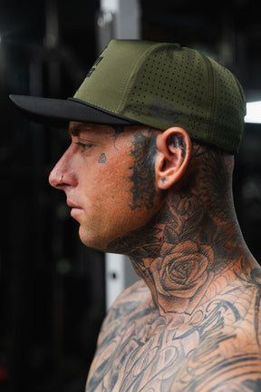 A shirtless man with extensive tattoos on his neck and face is shown in profile against a dark background, wearing the Tango Charlie Apparel Lift Heavy. Trust God. Hydro – OD Green Snapback Hat.