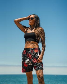 A woman with long dark hair and tattoos poses by the ocean under a clear blue sky, wearing sunglasses, a black crop top, and Relentless Betrayal Premium Mesh Shorts | 5- Thorn Amongst Roses.