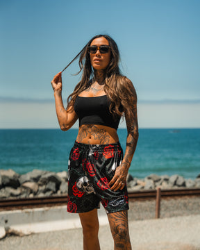 A woman with long hair, sunglasses, and tattoos stands by the ocean in a black crop top and Relentless Betrayals Premium Mesh Shorts | 5- Thorn Amongst Roses. Rocks, water, and blue sky form a striking backdrop.