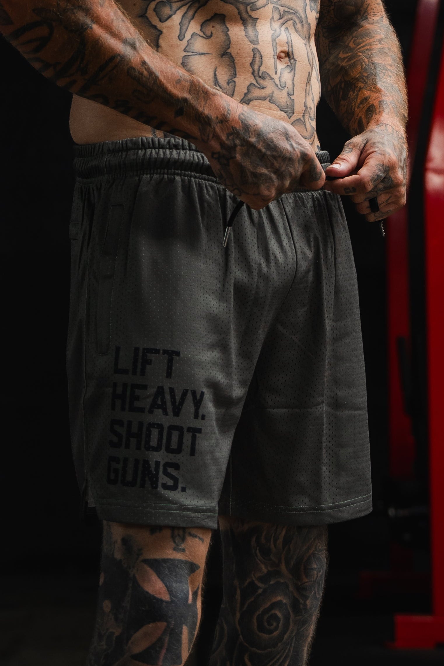 A tattooed person adjusts a pair of Tango Charlie Apparel Men's Mesh Lifestyle Shorts (5.5" - LHSG OD Green) featuring the text "LIFT HEAVY. SHOOT GUNS." Red gym equipment can be seen in the background.