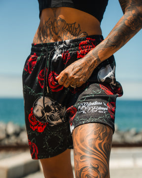 A tattooed person stands by the ocean on a sunny day, wearing Relentless Betrayals Premium Mesh Shorts | 5- Thorn Amongst Roses with red rose and skull designs, paired with a black crop top and hand resting on the waistband.