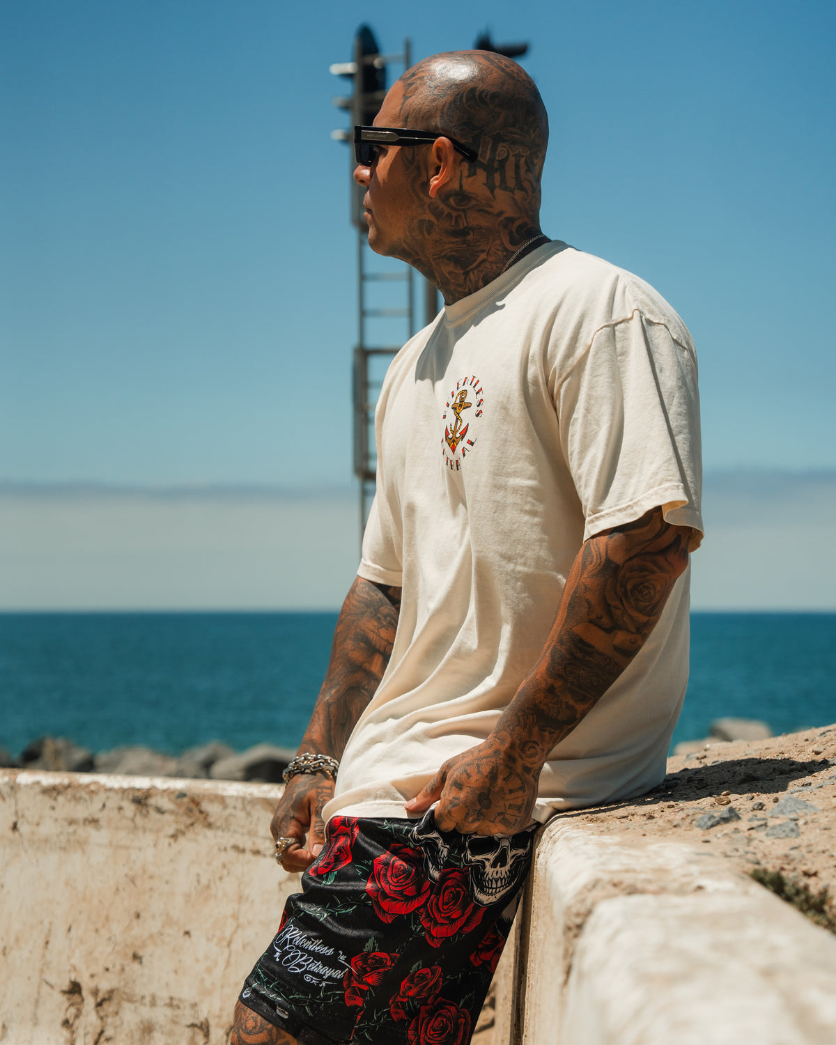 A tattooed man in sunglasses and a white T-shirt wears Relentless Betrayal’s Premium Mesh Shorts | 5- Thorn Amongst Roses as he leans on a concrete wall by the sea under a clear blue sky.