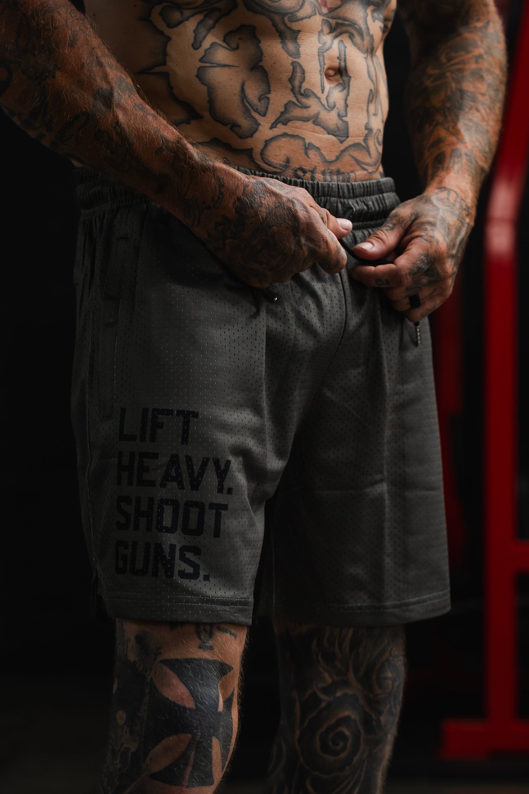 A tattooed man adjusts the waistband of the Tango Charlie Apparel Men's Mesh Lifestyle Shorts | 5.5"- LHSG OD Green, featuring "LIFT HEAVY. SHOOT GUNS." printed on the left leg, highlighting these versatile shorts designed for any active routine.