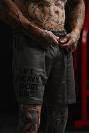 A tattooed man adjusts the waistband of the Tango Charlie Apparel Men's Mesh Lifestyle Shorts | 5.5"- LHSG OD Green, featuring "LIFT HEAVY. SHOOT GUNS." printed on the left leg, highlighting these versatile shorts designed for any active routine.