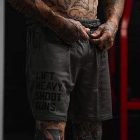 A tattooed person in a gym adjusts the drawstring on Tango Charlie Apparel Men's Mesh Lifestyle Shorts | 5.5"- LHSG OD Green, featuring "LIFT HEAVY. SHOOT GUNS." printed on the left leg.