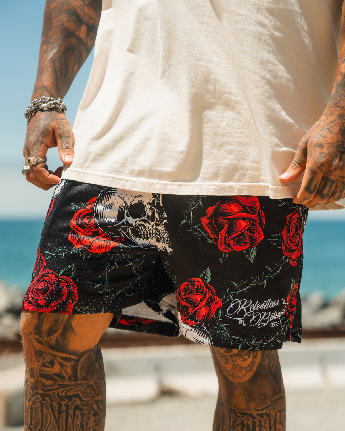 A person wearing Relentless Betrayal’s Premium Mesh Shorts | 5- Thorn Amongst Roses, featuring red rose and skull designs, stands by a body of water. Only their tattooed, jewelry-adorned lower half is visible.