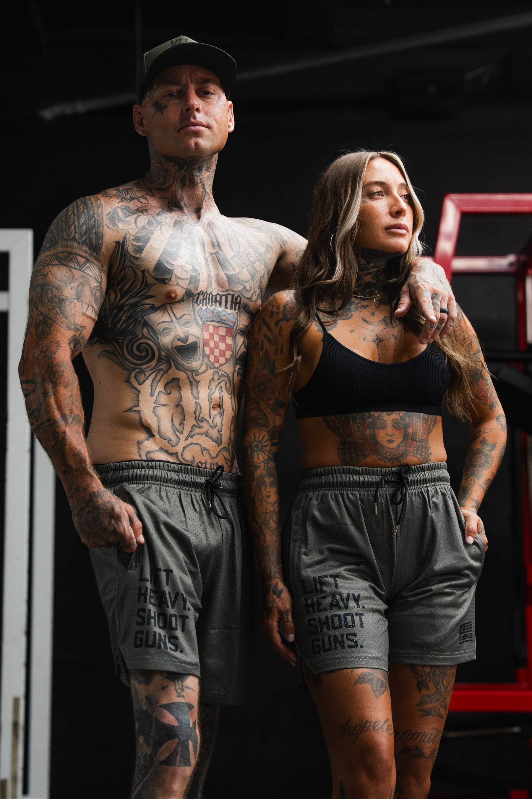 Two tattooed adults in gym attire stand close together, the man shirtless and the woman in a sports bra. Both are wearing Tango Charlie Apparel’s Men’s Mesh Lifestyle Shorts | 5.5"- LHSG OD Green, featuring “LIFT HEAVY. SHOOT GUNS.” graphics—stylish performance shorts designed for those who live with strength and intensity.