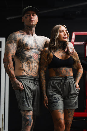 Two tattooed adults in gym attire stand close together, the man shirtless and the woman in a sports bra. Both are wearing Tango Charlie Apparel’s Men’s Mesh Lifestyle Shorts | 5.5"- LHSG OD Green, featuring “LIFT HEAVY. SHOOT GUNS.” graphics—stylish performance shorts designed for those who live with strength and intensity.