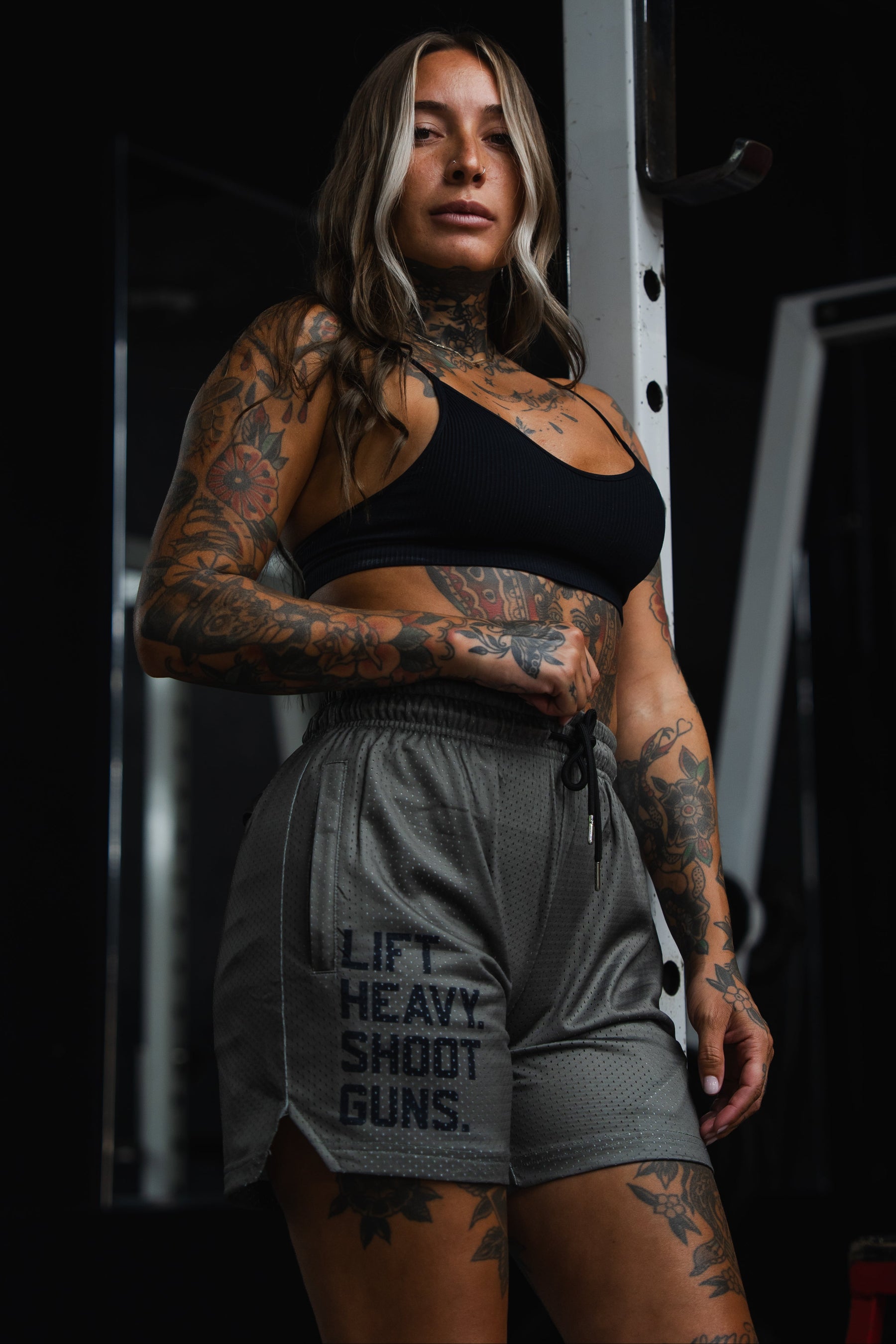 A tattooed woman stands in a gym wearing Tango Charlie Apparel's Men's Mesh Lifestyle Shorts | 5.5"- LHSG OD Green, featuring the phrase “LIFT HEAVY. SHOOT GUNS.” on the gray workout shorts and a black sports bra, looking at the camera with gym equipment visible in the background.