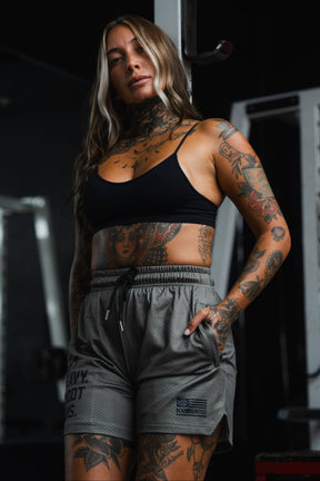 Woman with long hair and visible tattoos poses in a gym, wearing Tango Charlie Apparel’s Men's Mesh Lifestyle Shorts in 5.5" LHSG OD Green, with her hands in her pockets—perfect for showcasing effortless style in these versatile shorts.