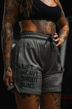 A tattooed individual wearing a black crop top and Tango Charlie Apparel Men's Mesh Lifestyle Shorts | 5.5"- LHSG OD Green, featuring "LIFT HEAVY. SHOOT GUNS." printed on the left leg, stands with one hand on their hip.