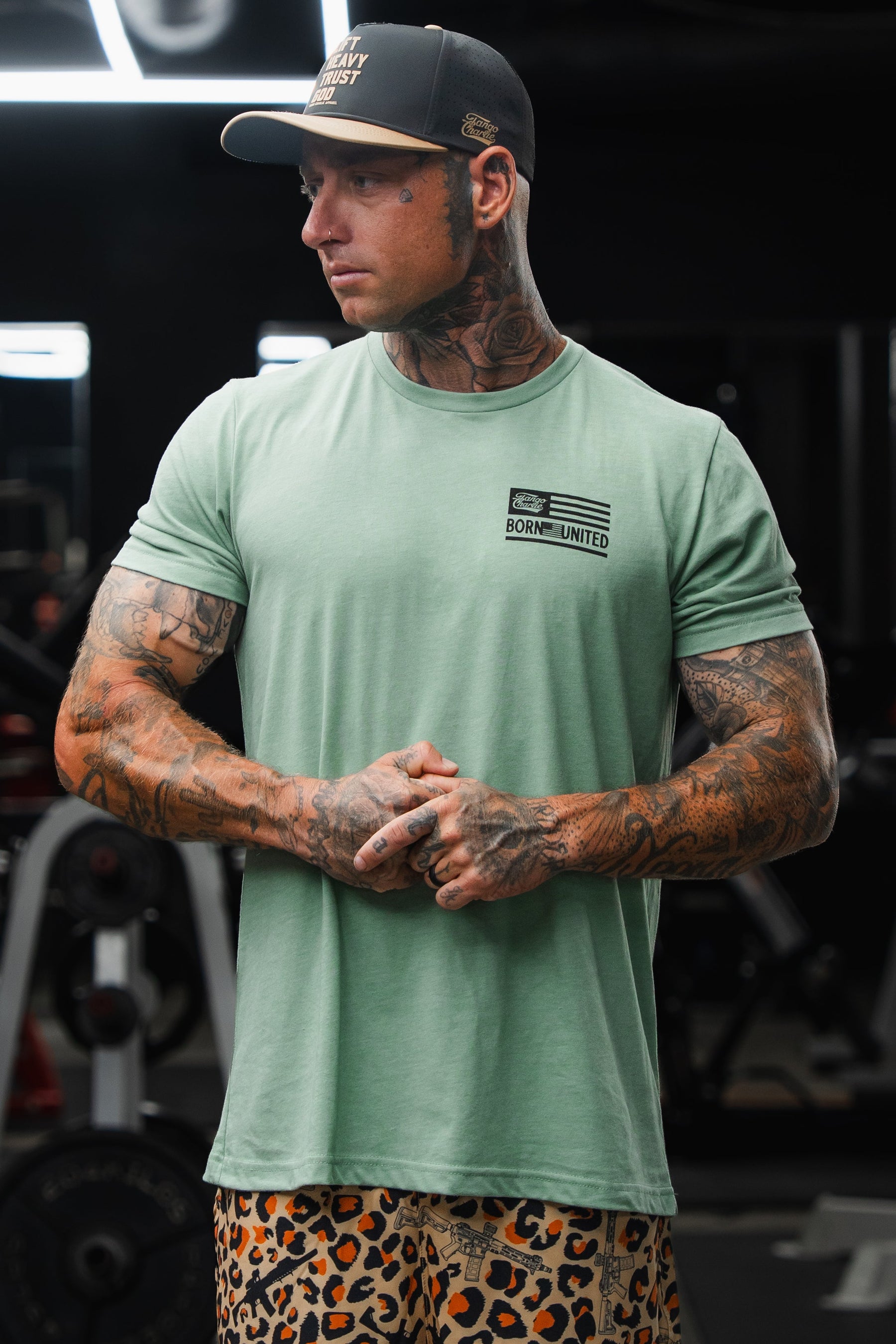 A tattooed man stands in a gym, facing sideways with his hands clasped, wearing the Loaded Leopard LHSG - Seafoam Green Tee by Tango Charlie Apparel and leopard print shorts—capturing the essence of high-intensity fitness style.
