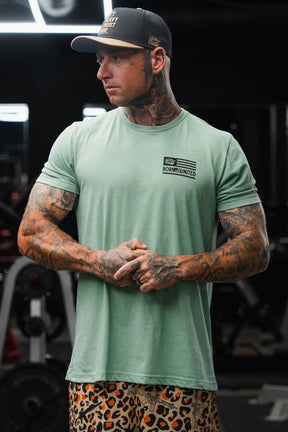 A tattooed man stands in a gym, facing sideways with his hands clasped, wearing the Loaded Leopard LHSG - Seafoam Green Tee by Tango Charlie Apparel and leopard print shorts—capturing the essence of high-intensity fitness style.