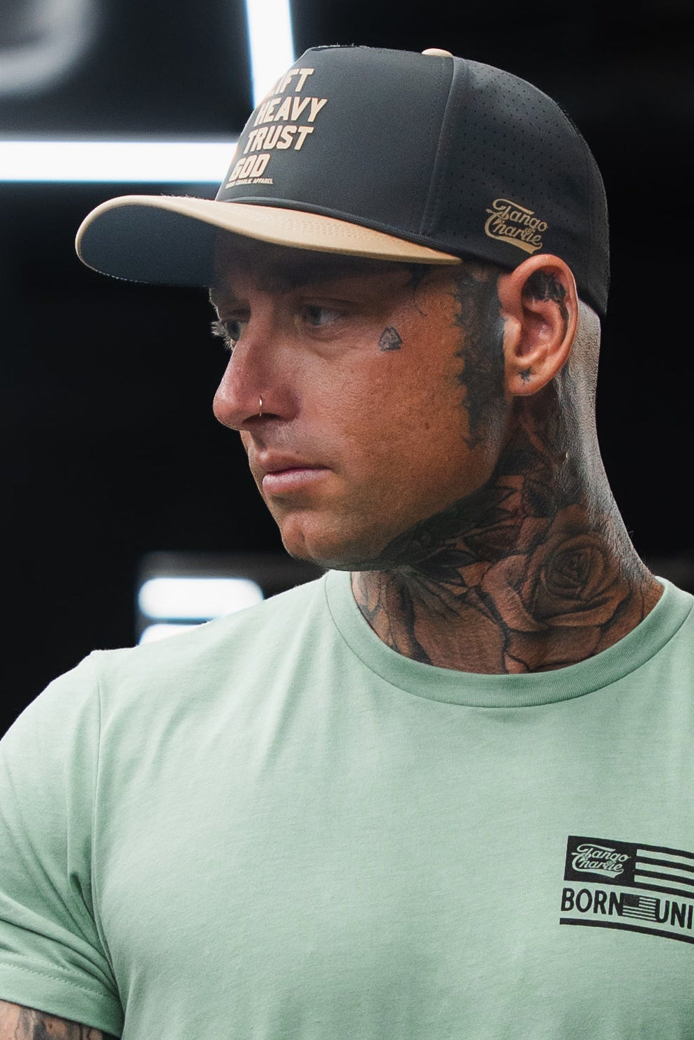Tattooed man wearing a green T-shirt and a Tango Charlie Apparel Lift Heavy. Trust God. Hydro black snapback hat, looking to the side.