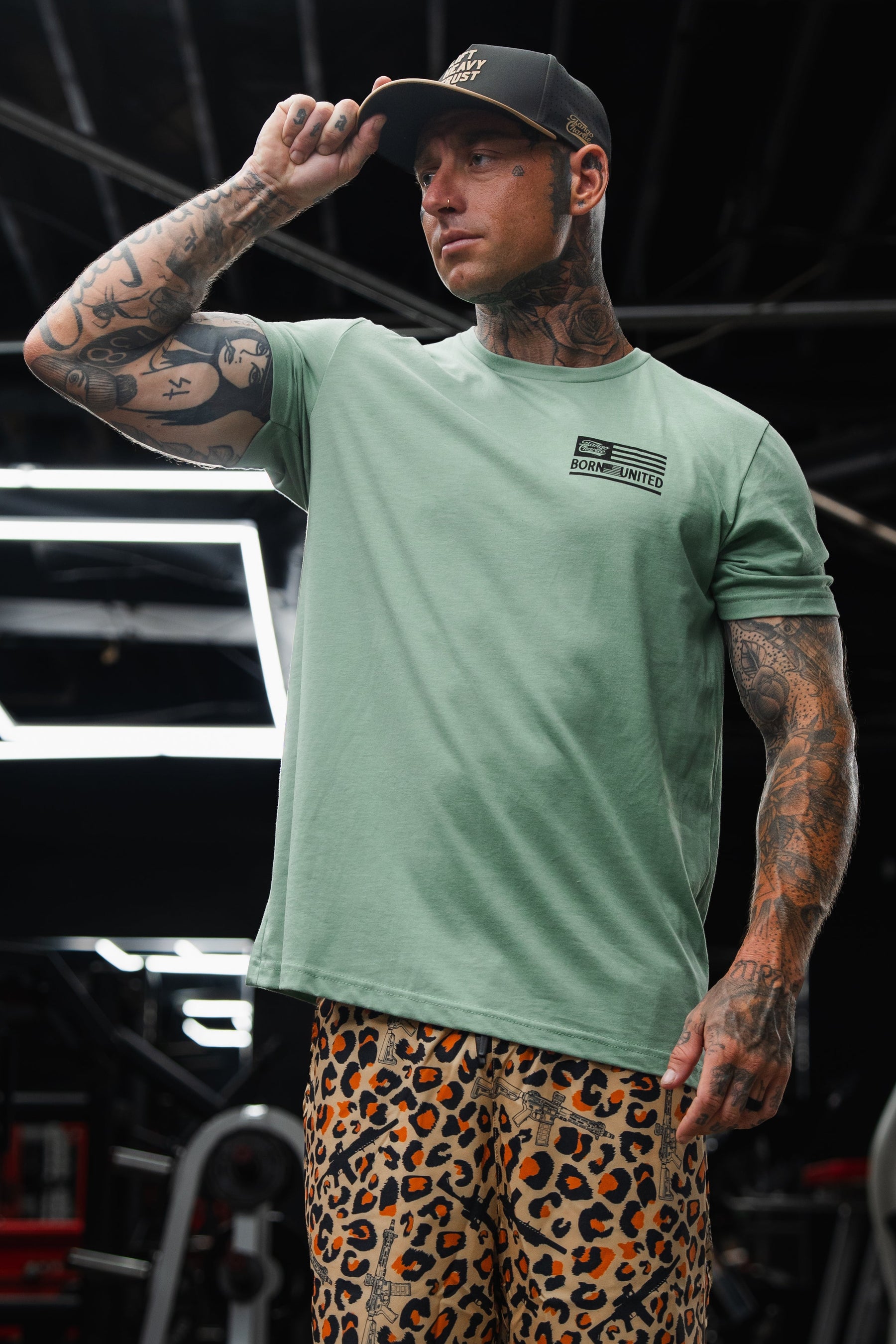 A tattooed man in a Tango Charlie Apparel Loaded Leopard LHSG - Seafoam Green Tee and leopard print pants adjusts his black cap while standing in a gym, highlighting his high-intensity fitness gear.