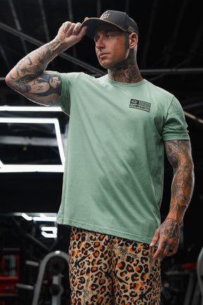 A tattooed man in a Tango Charlie Apparel Loaded Leopard LHSG - Seafoam Green Tee and leopard print pants adjusts his black cap while standing in a gym, highlighting his high-intensity fitness gear.