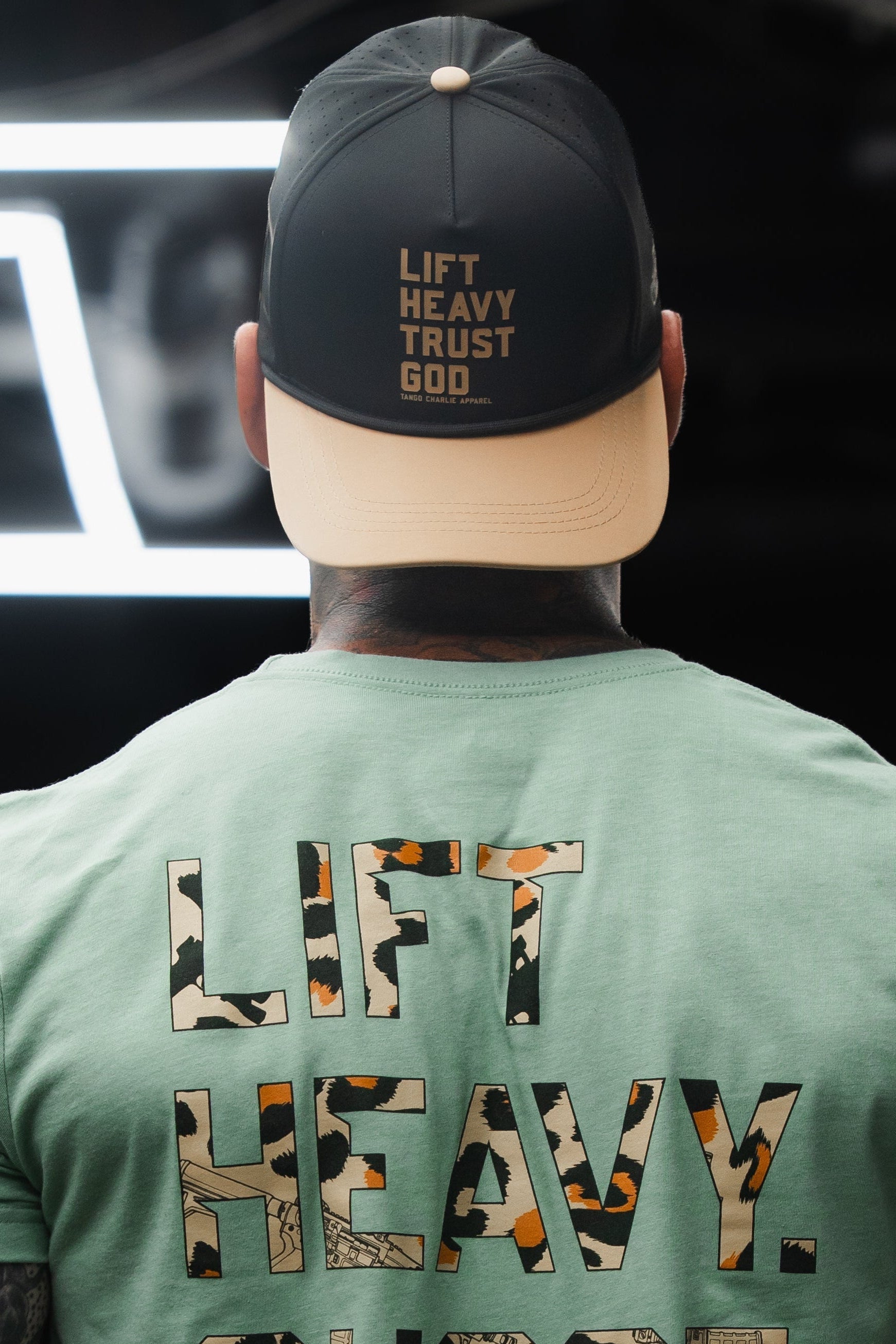 A person, shown from behind, is wearing a Tango Charlie Apparel Lift Heavy. Trust God. Hydro Black Snapback Hat and a matching t-shirt, both featuring the phrase “Lift Heavy, Trust God.”.