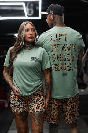 Two tattooed individuals, both wearing seafoam green Loaded Leopard LHSG tees from Tango Charlie Apparel and leopard print shorts, stand confidently in a gym surrounded by high-intensity fitness gear. The man’s shirt features the bold “LIFT HEAVY. SHOOT GUNS.” slogan, representing their dedication to both workouts and Second Amendment pride. Gym equipment fills the background, creating a striking atmosphere of strength and unity.
